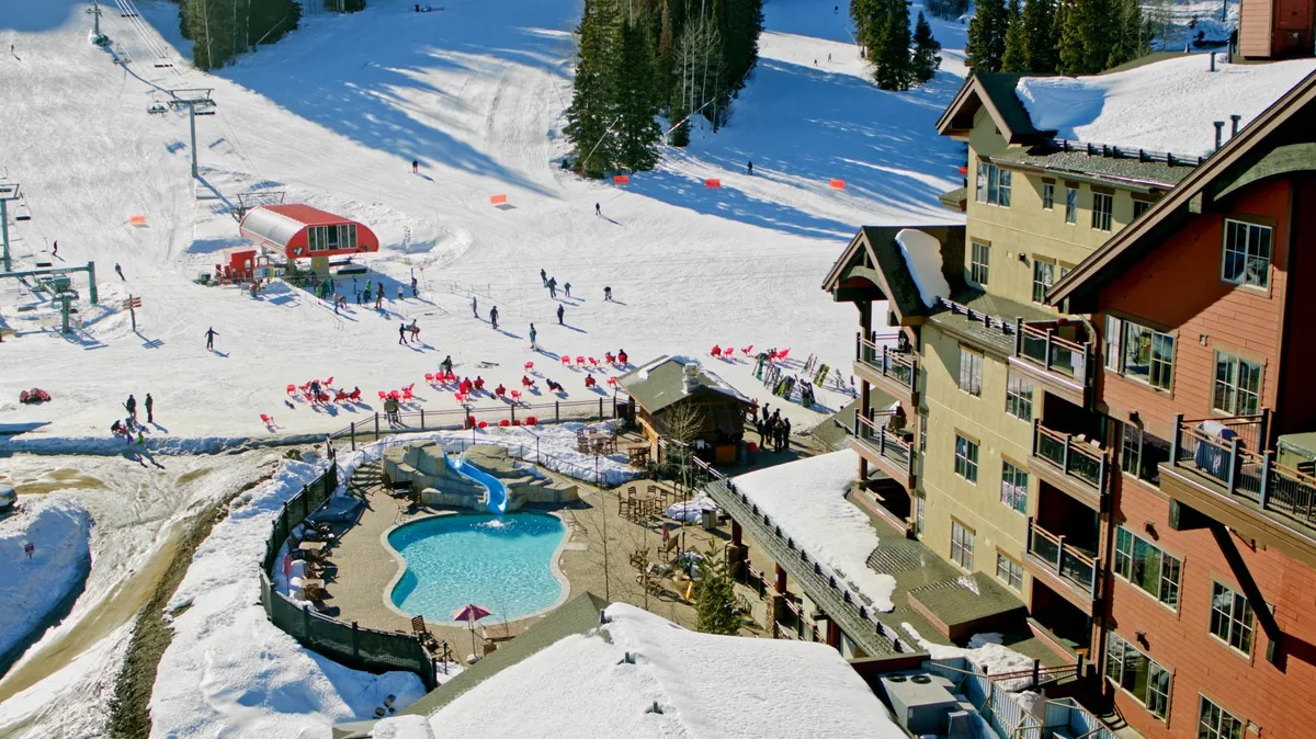 A wide exterior shot showcasing the Purgatory Townhomes complex set against the backdrop of snowy ski slopes, a bustling pool area, and the ski lift nearby. The architecture blends with the natural mountain scenery, evoking a sense of adventure and possibility.