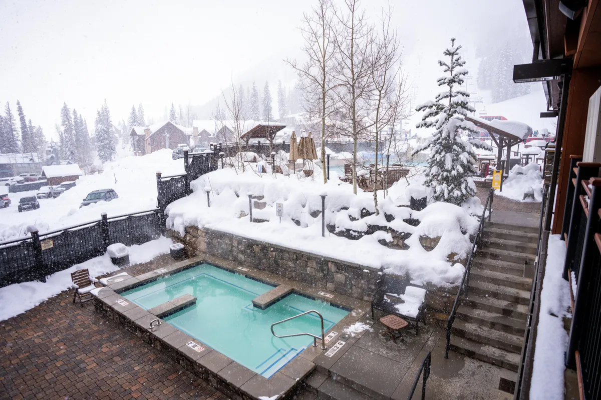 A serene outdoor hot tub steams amid a snowy landscape, surrounded by trees blanketed in snow. People relax in the warmth, some sipping hot drinks and socializing while gazing at the wintry scenery. A staircase leads up to the property, emphasizing easy access after a day on the slopes.