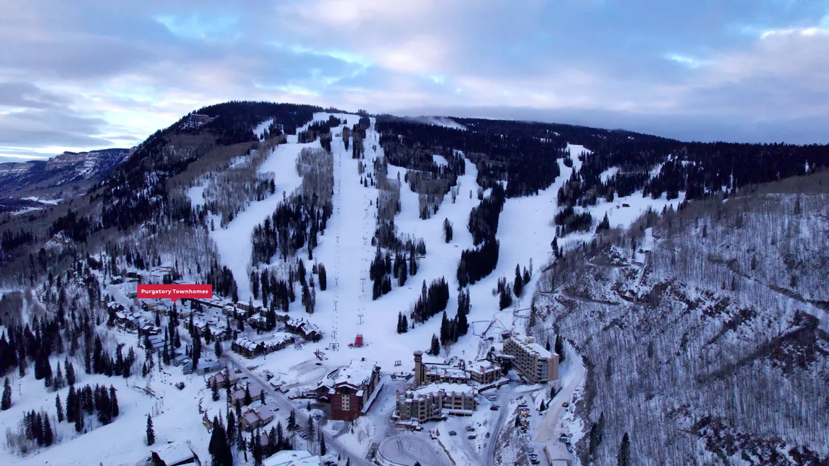 A panoramic view reveals snow-dusted townhomes set against forested slopes and ready ski trails. The early morning or dusk lighting suggests a peaceful transition between adventure on the slopes and cozy evenings in, with the inviting proximity of home clear in the frame.
