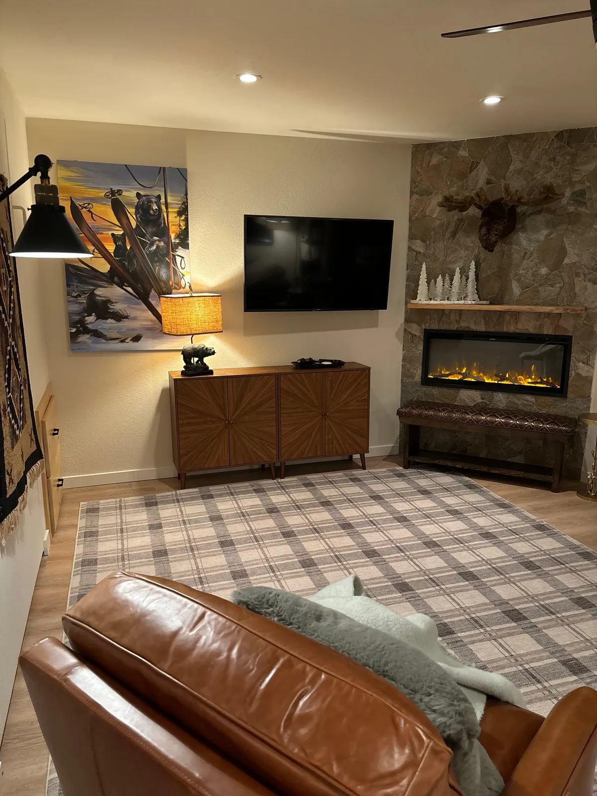 A cozy living area featuring a bear painting above a wooden cabinet, a large television, a leather sofa, and a glowing fireplace. The decor is inviting, with warm tones and comfortable furnishings that evoke the ambiance of a mountain retreat.