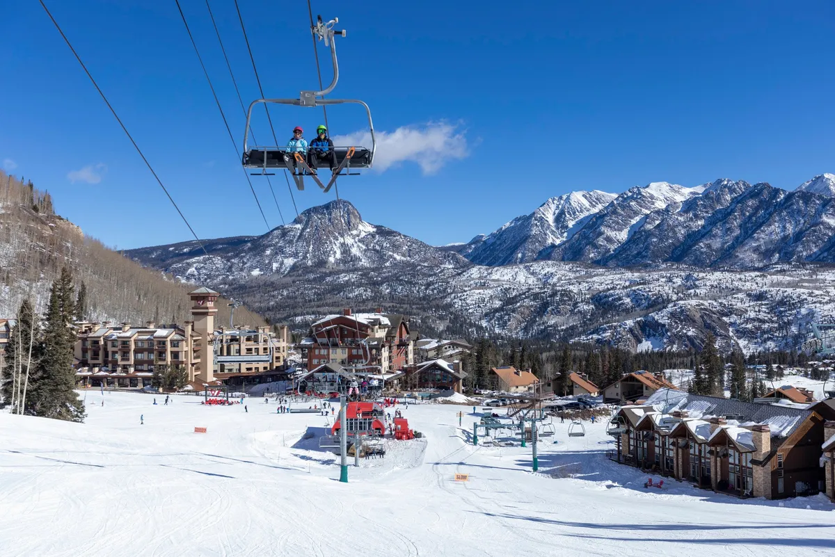 A sweeping exterior view captures the heart of winter activity at Purgatory Resort: a bustling ski lift transports skiers and snowboarders over snowy slopes, with the ski resort buildings nestled below. People are actively skiing and snowboarding, while the snow-covered mountains create a dramatic, energetic backdrop.
