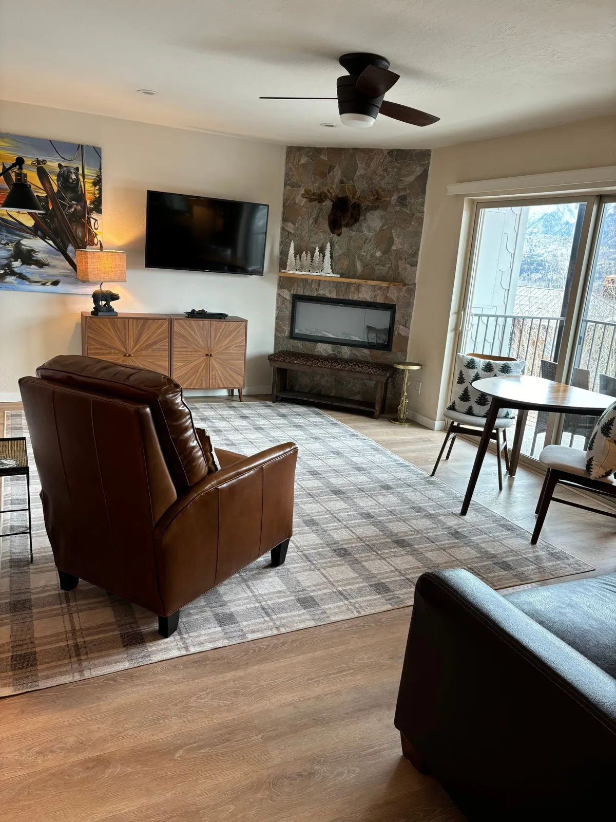 A cozy, inviting living space featuring a plush leather armchair, a beautiful stone fireplace, tasteful wildlife artwork, a modern television, and a small dining table—all arranged to create a warm, intimate atmosphere perfect for relaxing or unwinding after a day on the slopes.