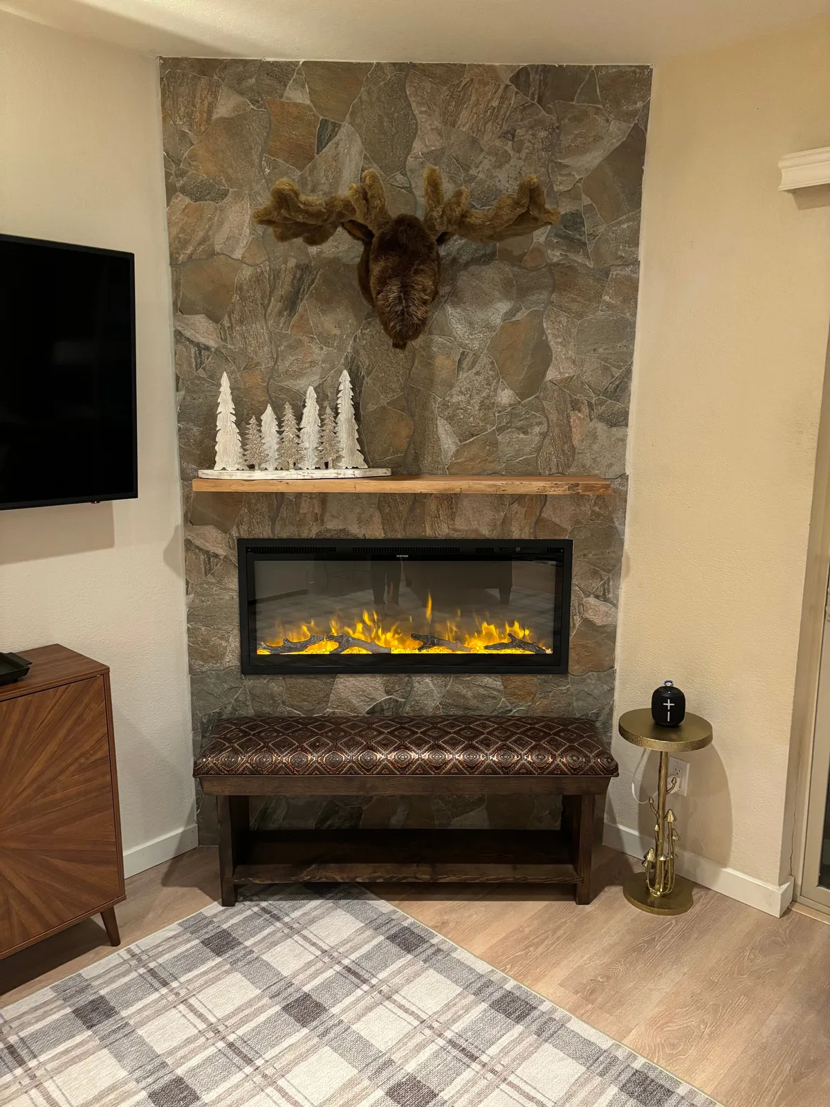 An inviting fireplace scene decorated with a moose head, decorative trees, a bench, and a plaid rug. The arrangement suggests a welcoming environment perfect for socializing or relaxing after a day out in the village.