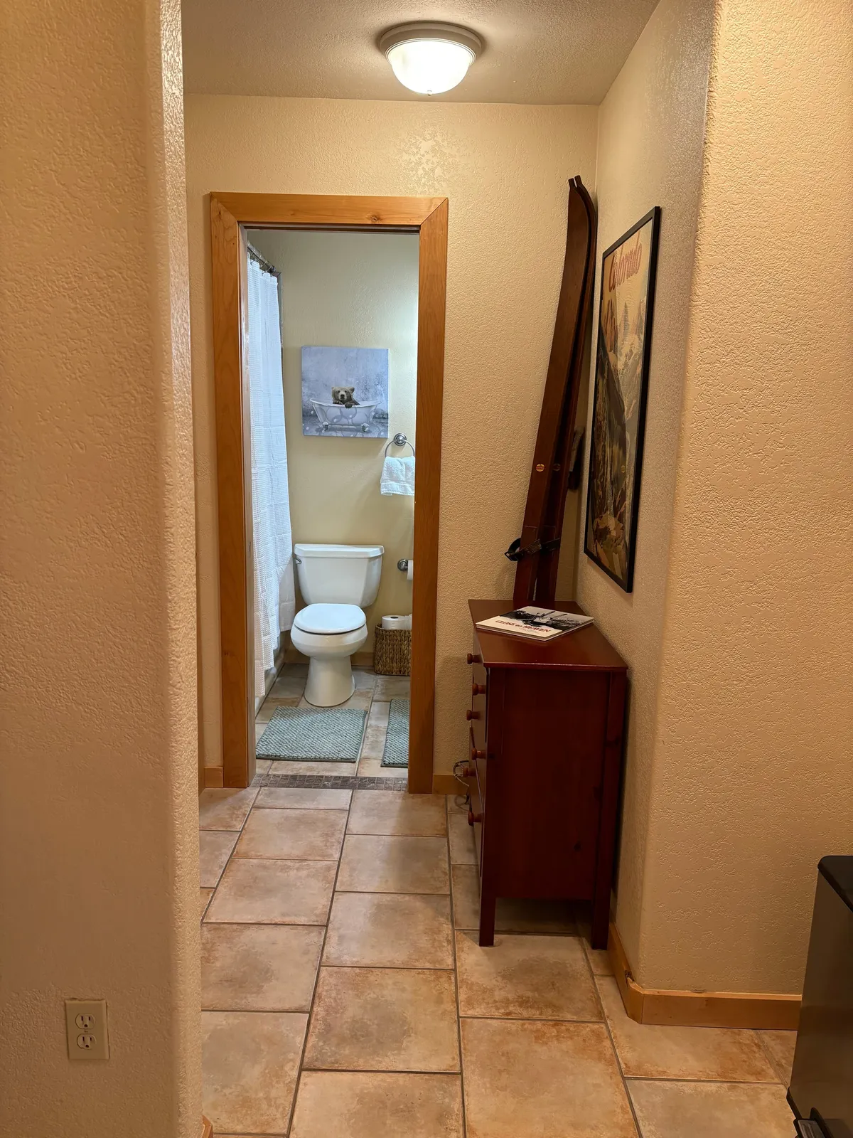 This image captures the entrance to a bathroom, emphasizing the transition from active pursuits (suggested by the visible ski equipment) to relaxation. A cozy wooden cabinet and framed artwork enhance the sense of comfort and warmth, while the open door hints at the welcoming bathroom within.