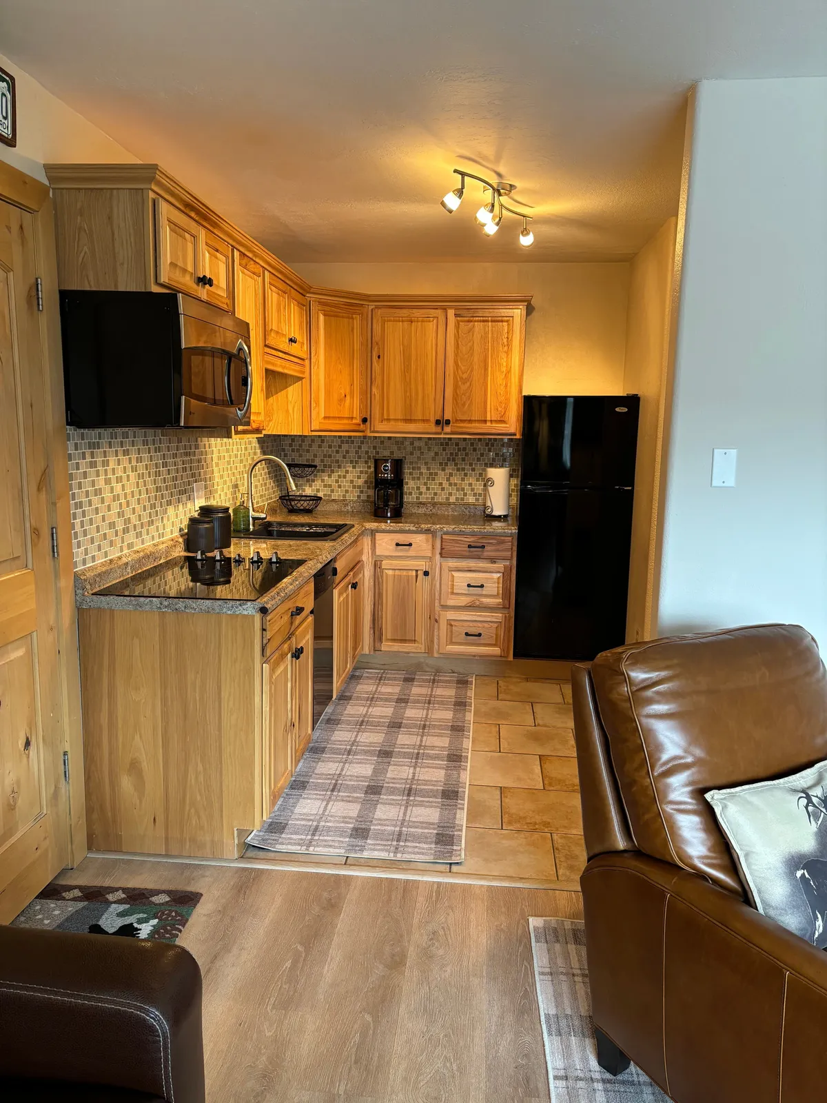 This image showcases a cozy kitchenette featuring a granite countertop, wooden cabinetry, and black appliances, with a plaid rug adding warmth to the space. The kitchen opens onto a living area with a leather sofa, creating a seamless, inviting environment for cooking and conversation.