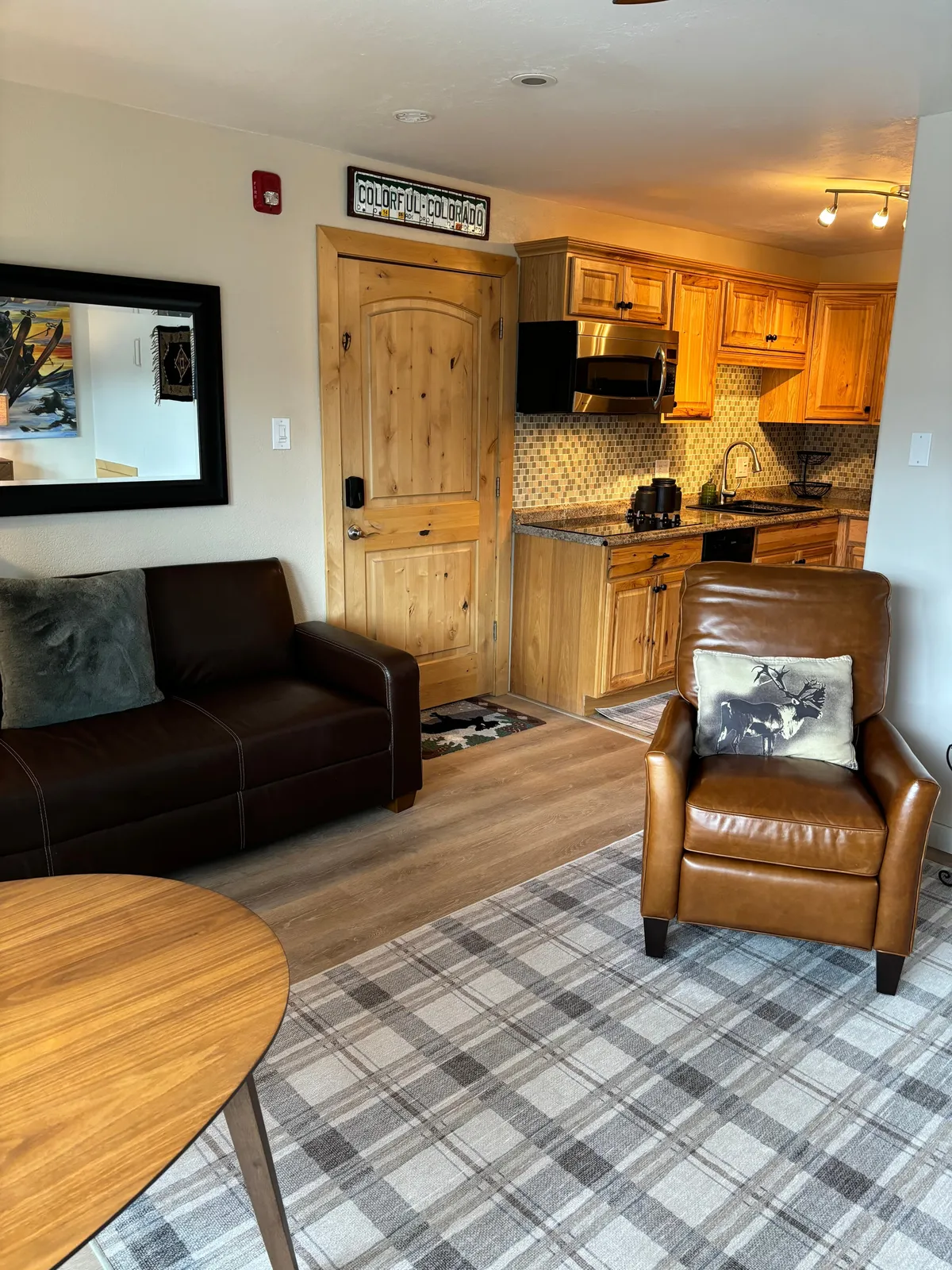 This image showcases an open-plan living space featuring a brown leather sofa, a plaid rug, a cozy leather armchair, and a view into the kitchen area. The setting is warm and inviting, with functional furnishings arranged for both comfort and efficiency. The wooden door and soft lighting further enhance the intimate ambiance.
