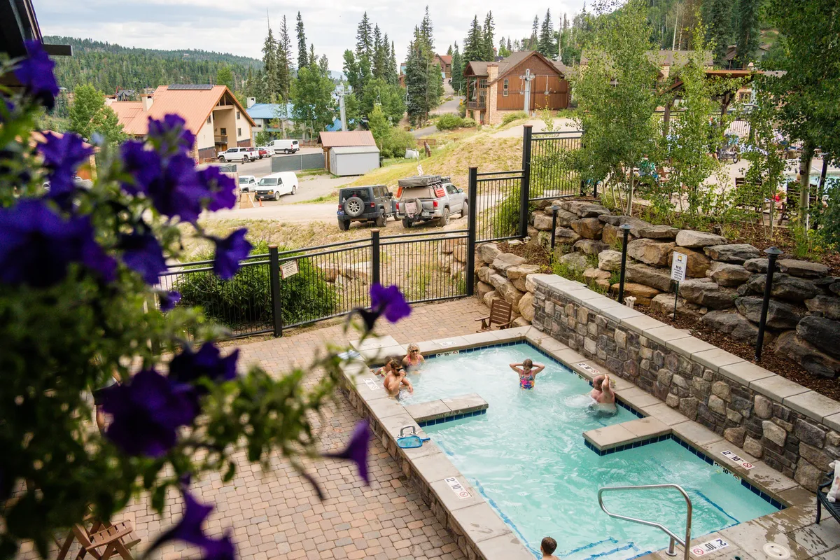 An inviting hot tub surrounded by colorful flowers and trees, with mountain views and residential buildings visible in the distance. The setting is tranquil, perfect for unwinding after a day outdoors, and connects the natural setting with relaxation amenities.